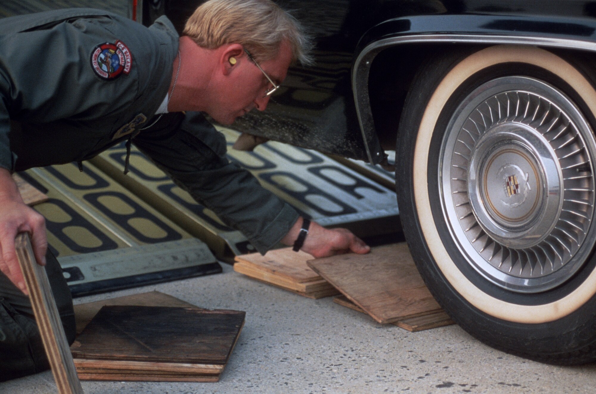 A man in uniform is placing a piece of wood near the front of a car tire so it can drive up the aircraft ramp with ease.