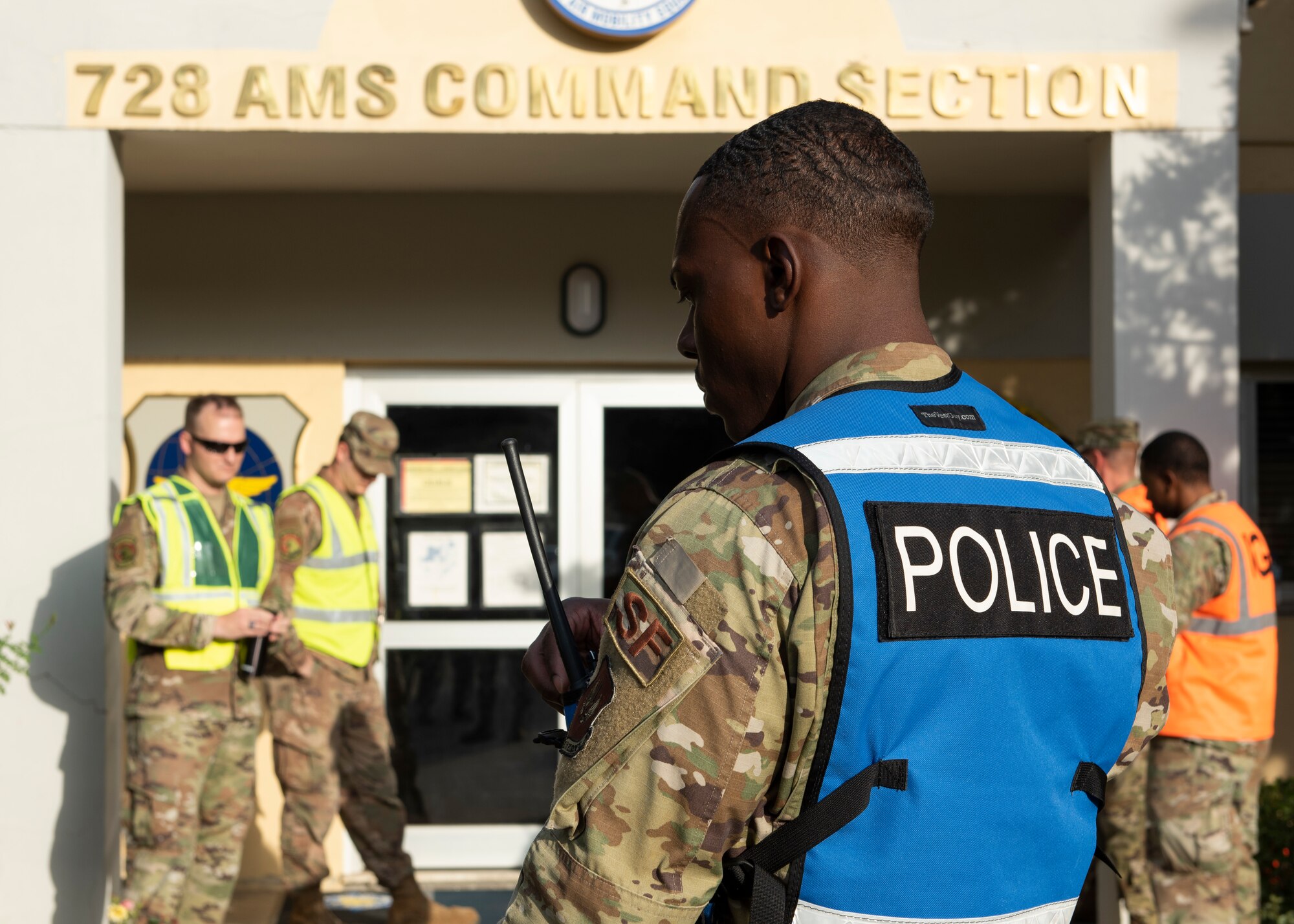 A blue vested security forces Airman checks his radio while several yellow vested Airmen wait in-front of a building entrance in the background.