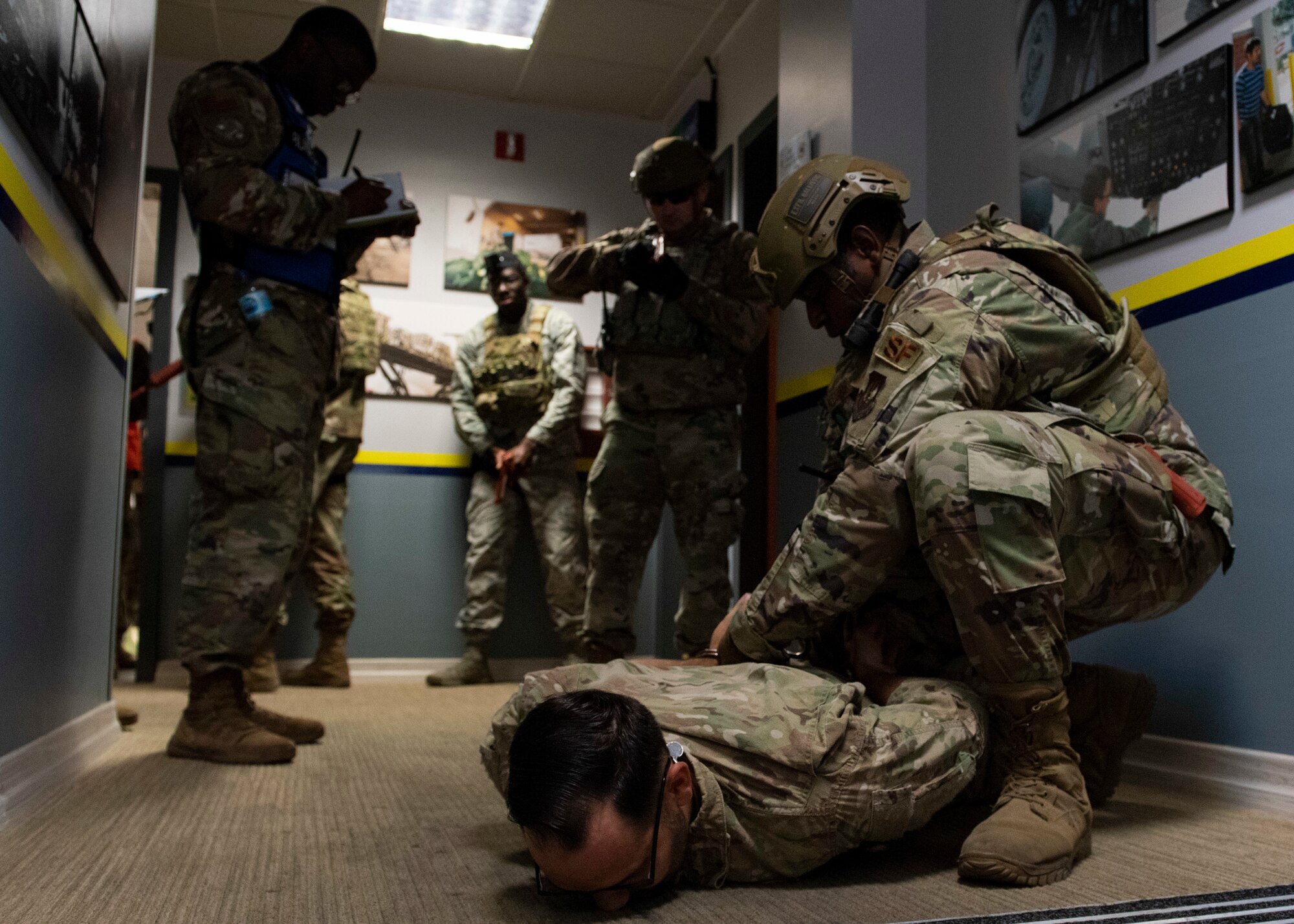 A handcuffed suspect lies face-down on the floor of an office building hallway, surrounded by five security forces Airmen, one of which kneels on top of the suspect to restrain him.
