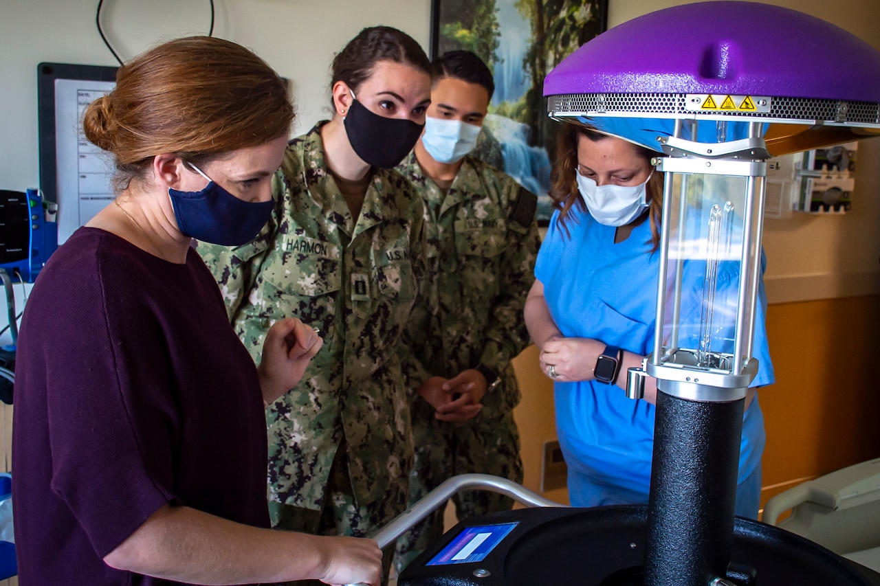 A woman wearing hospital scrubs shows a robot using ultraviolet light disinfection to another woman wearing scrubs and two people wearing military uniforms; all are wearing face masks.