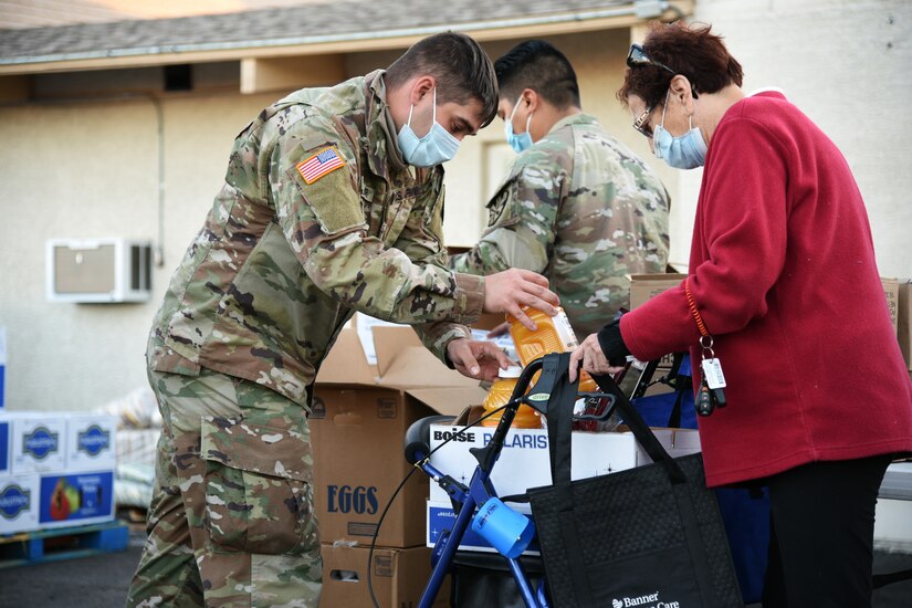 A service member wearing a face mask helps fill a grocery cart and bags for a senior citizen using a walker.