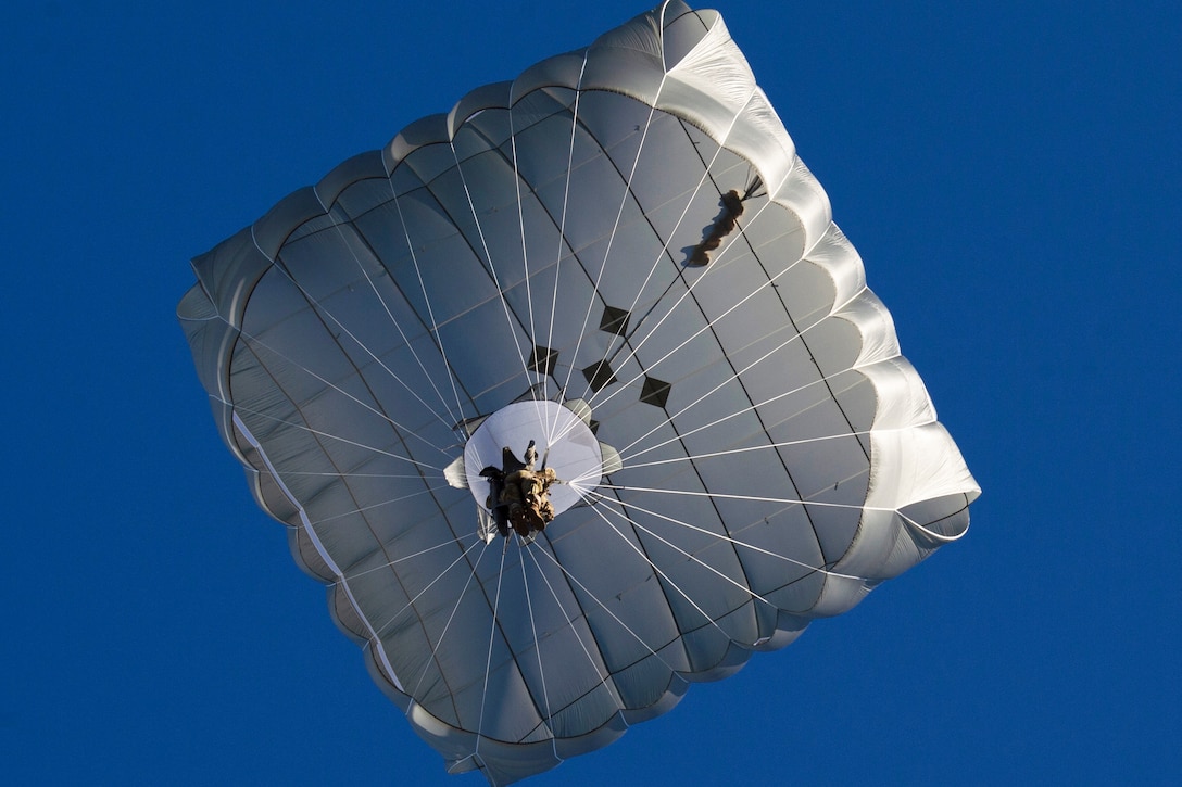 A soldier holds onto a large parachute as he floats downwards with blue sky around him.