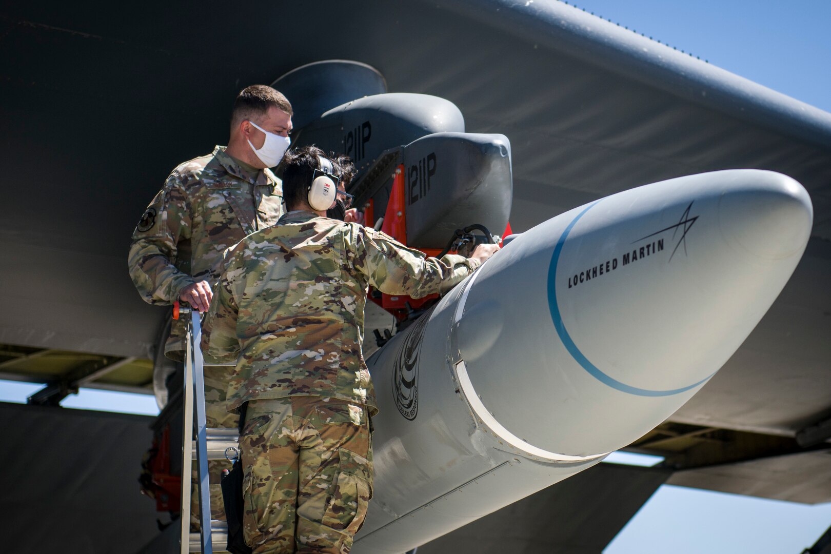 Two men in military uniforms stand on a ladder at the nose of an airplane.
