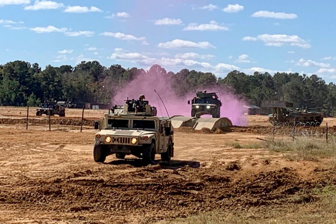 Army Humvees and other military vehicles drive on a dirt field, with purple smoke rising in the distance.