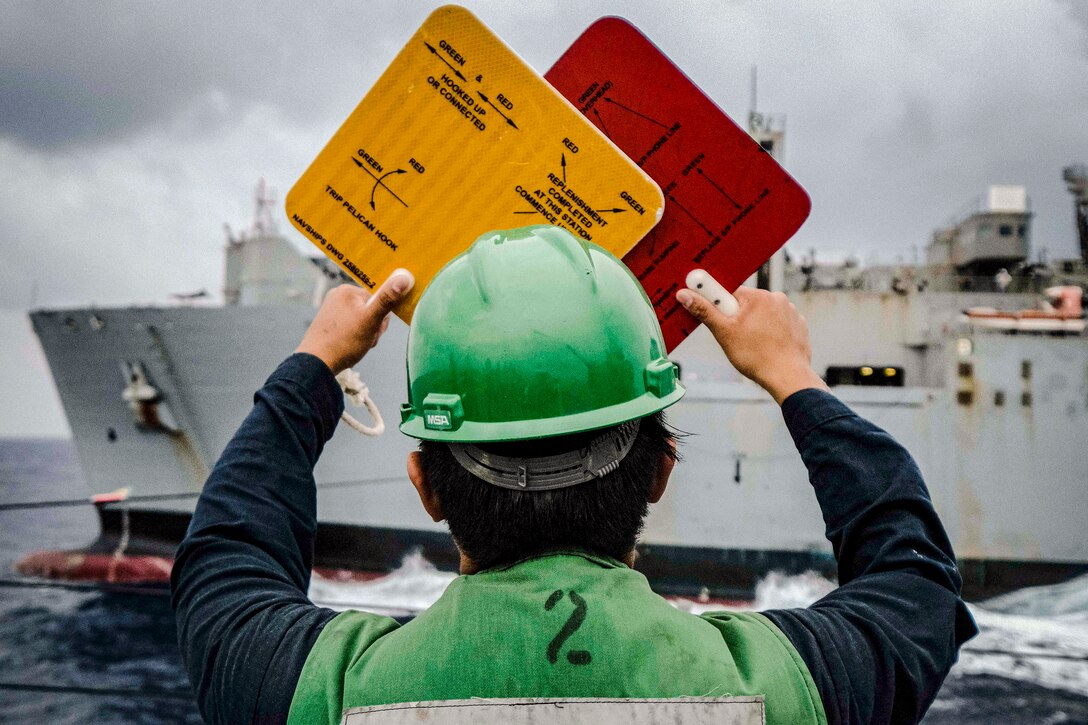 A sailor in green aboard a ship's deck facing another ship holds up a yellow square and red square.