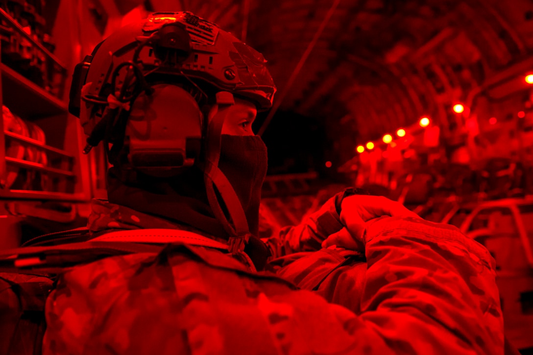 An airman, shown in profile and illuminated by red light, sits on an aircraft and holds a bundle on his lap.