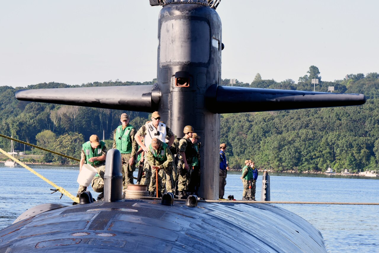 Sailors stand on top of a submarine while one of them holds a line and another holds a bucket.