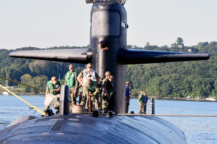 Sailors stand on top of a submarine while one of them holds a line and another holds a bucket.