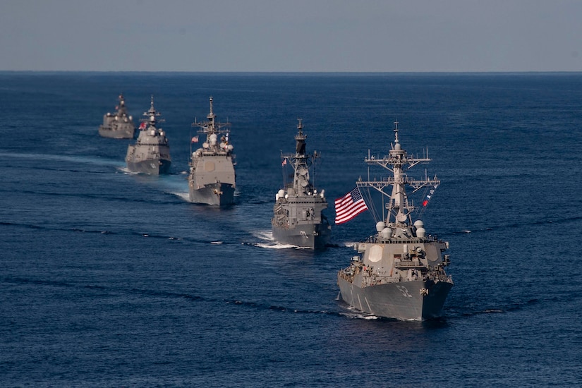 Five ships, the first carrying an American flag, sail through the ocean.