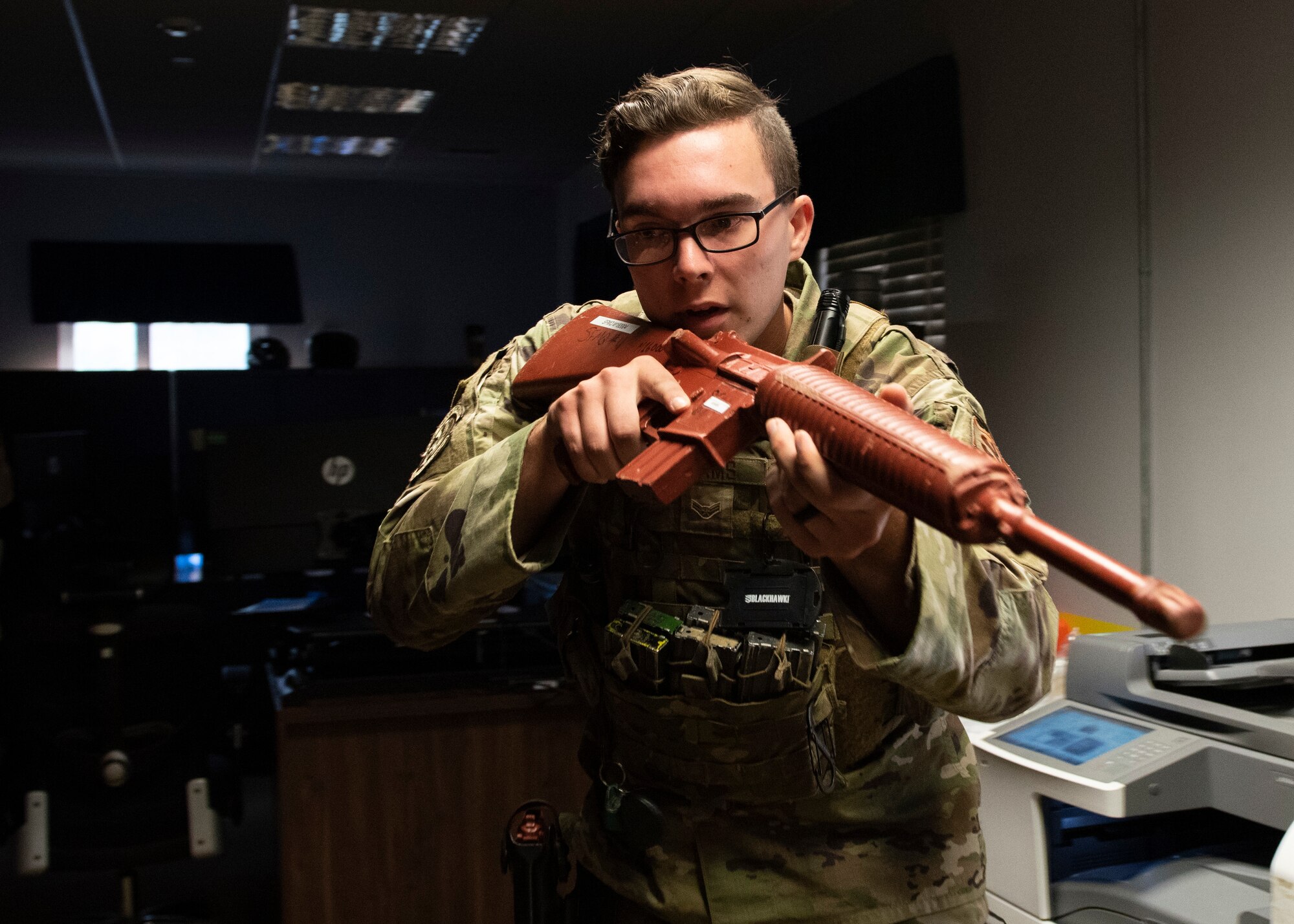 A security forces Airman holds up a red, practice M-16 rifle while slowly walking through a dark office space.
