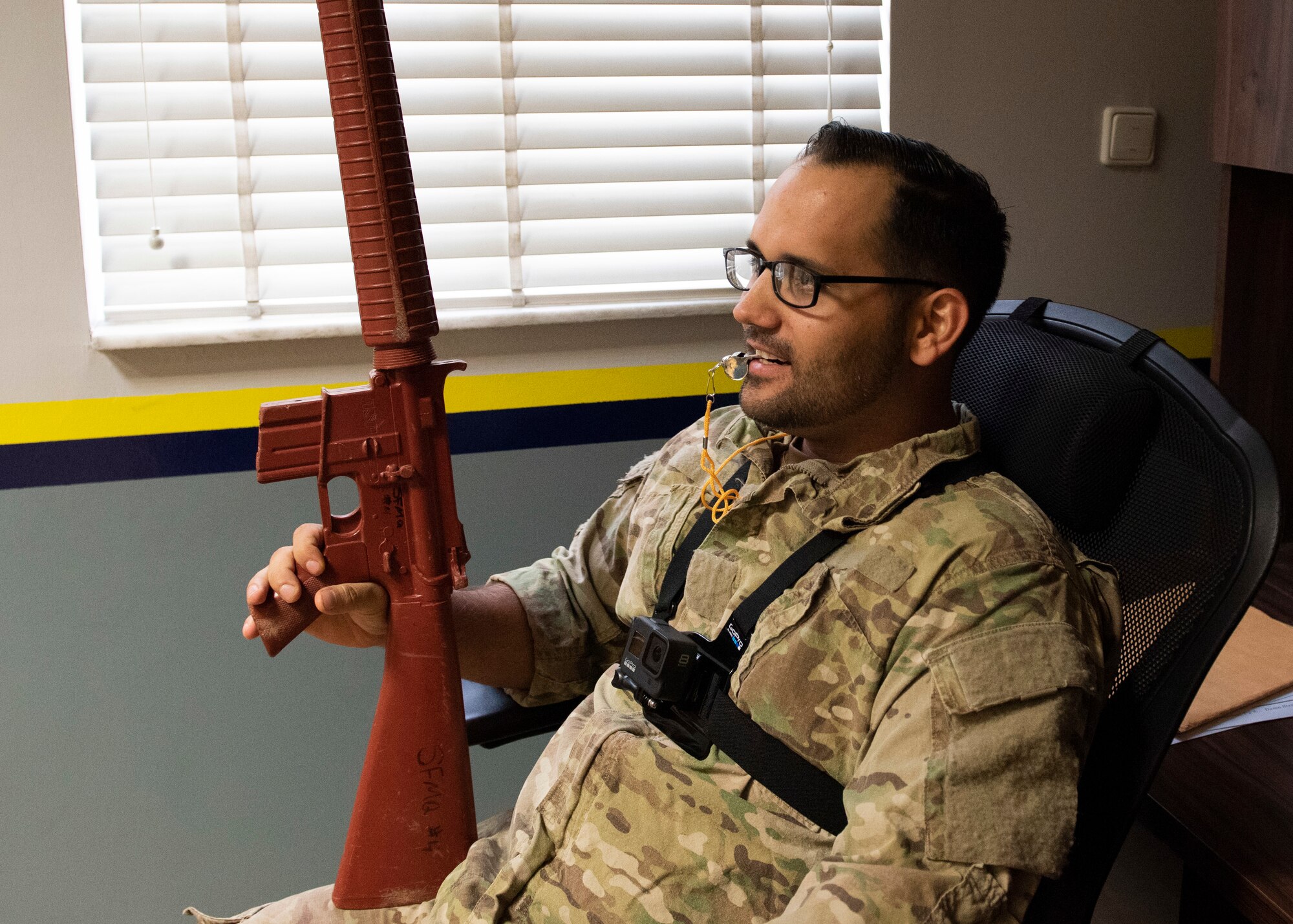 An Airman sits in an office chair while holding a red training M-16 rifle in one hand and has a whistle in his mouth.