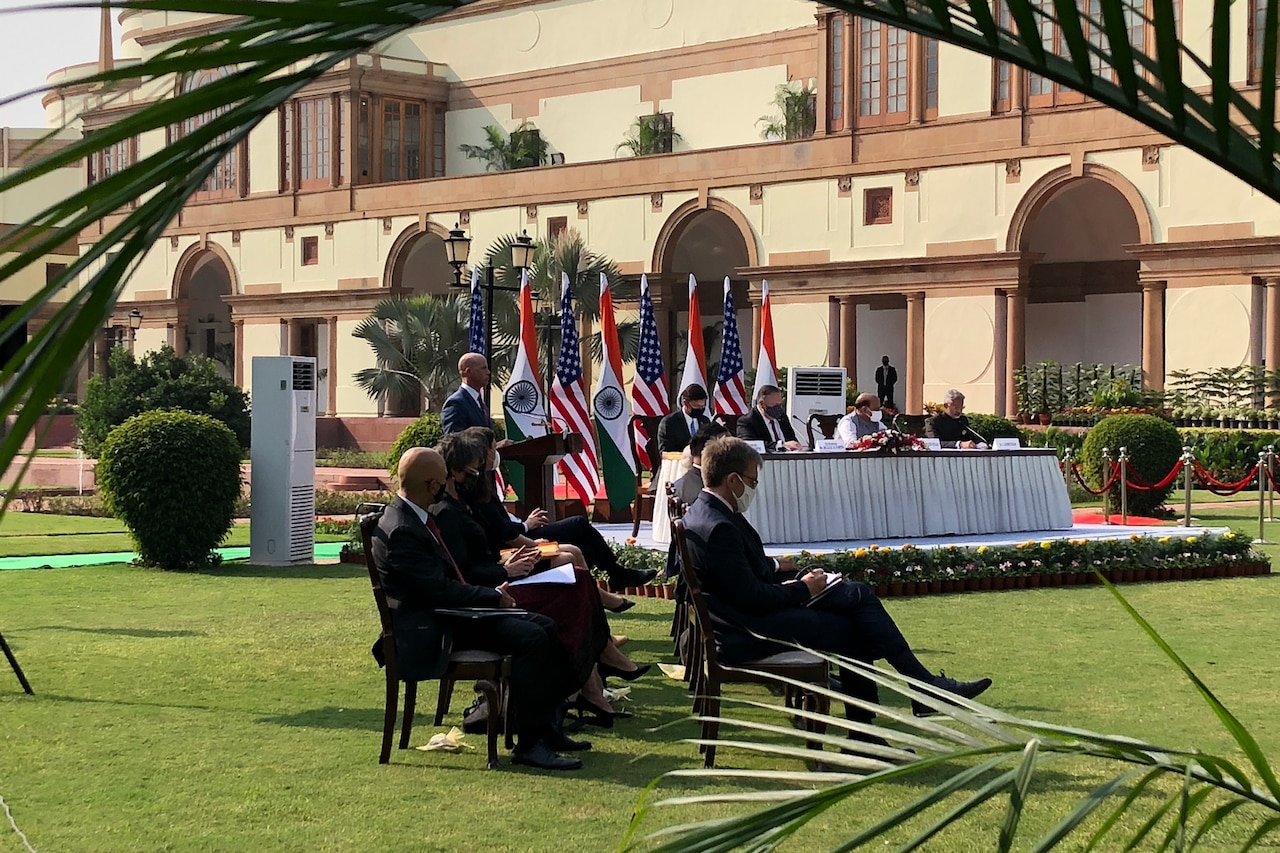 Four men sit at a long table in front of U.S. and Indian flags.