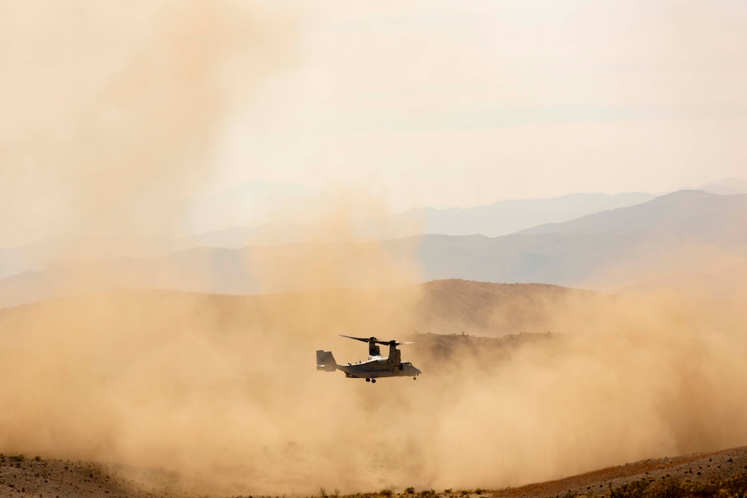 A military aircraft flies above a desert terrain as dust lifts from the ground.