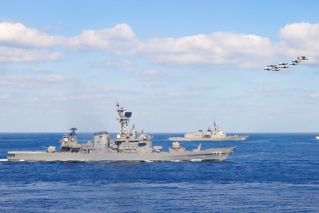 U.S. Navy, U.S. Marine Corps, Japan Maritime Self-Defense Force and Japan Air Self Defense Force aircraft fly in formation above Japanese and Canadian vessels in a body of water.