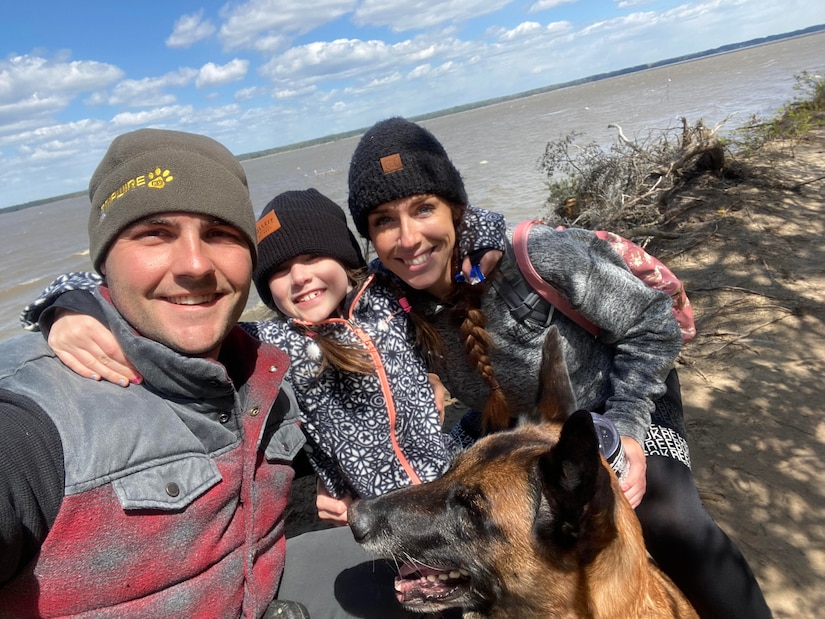 A man, woman and young girl huddle together with a dog near a body of water.