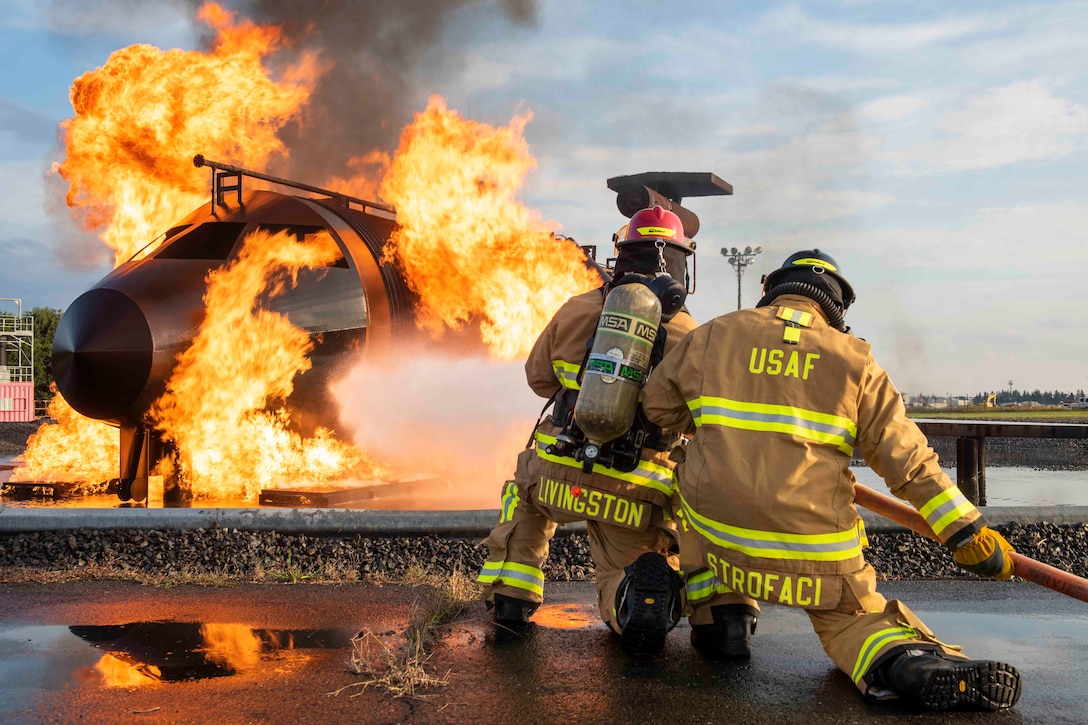 Two airmen use a hose to spray water onto an aircraft fire.