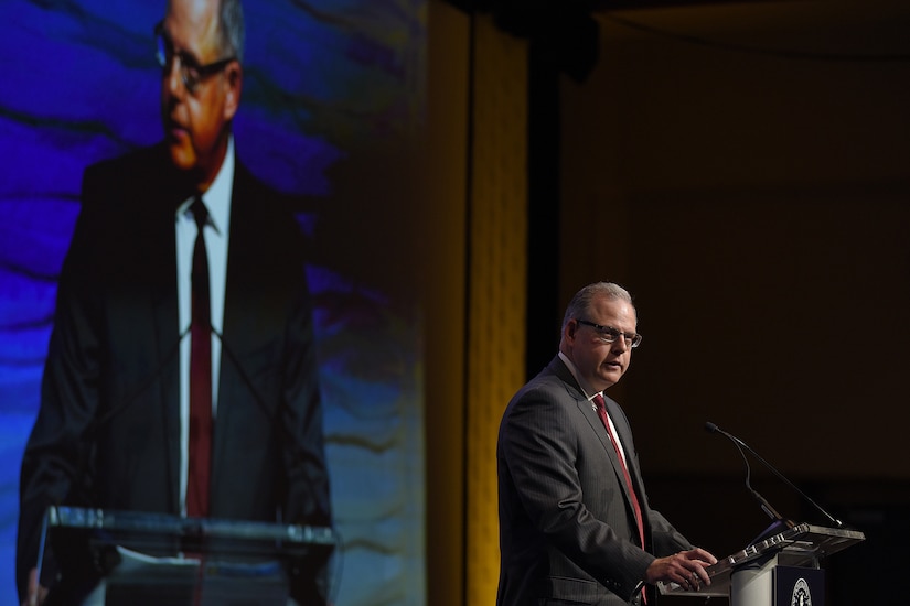 A man stands behind a lectern. Behind him, he is seen on a large video screen.