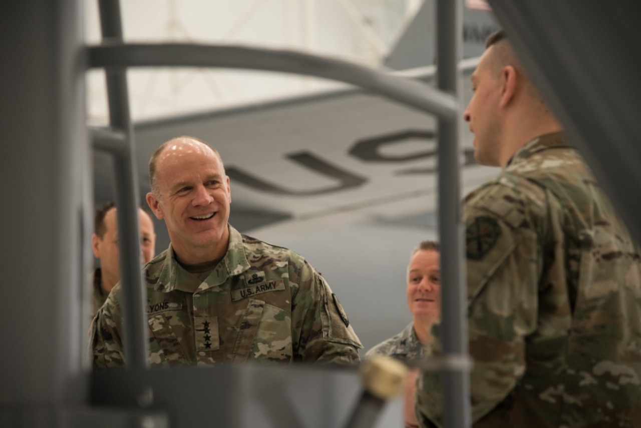 An army general smiles. His face is framed by military equipment in the foreground. Other soldiers stand near the general.