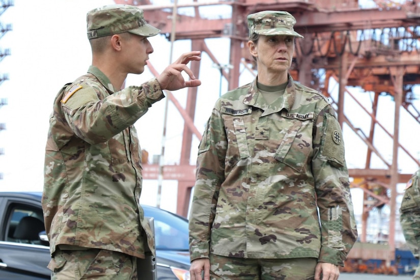 Two soldiers stand in front of a black car. One, a high-ranking officer, is looking away from the other as he gestures to something in the distance.