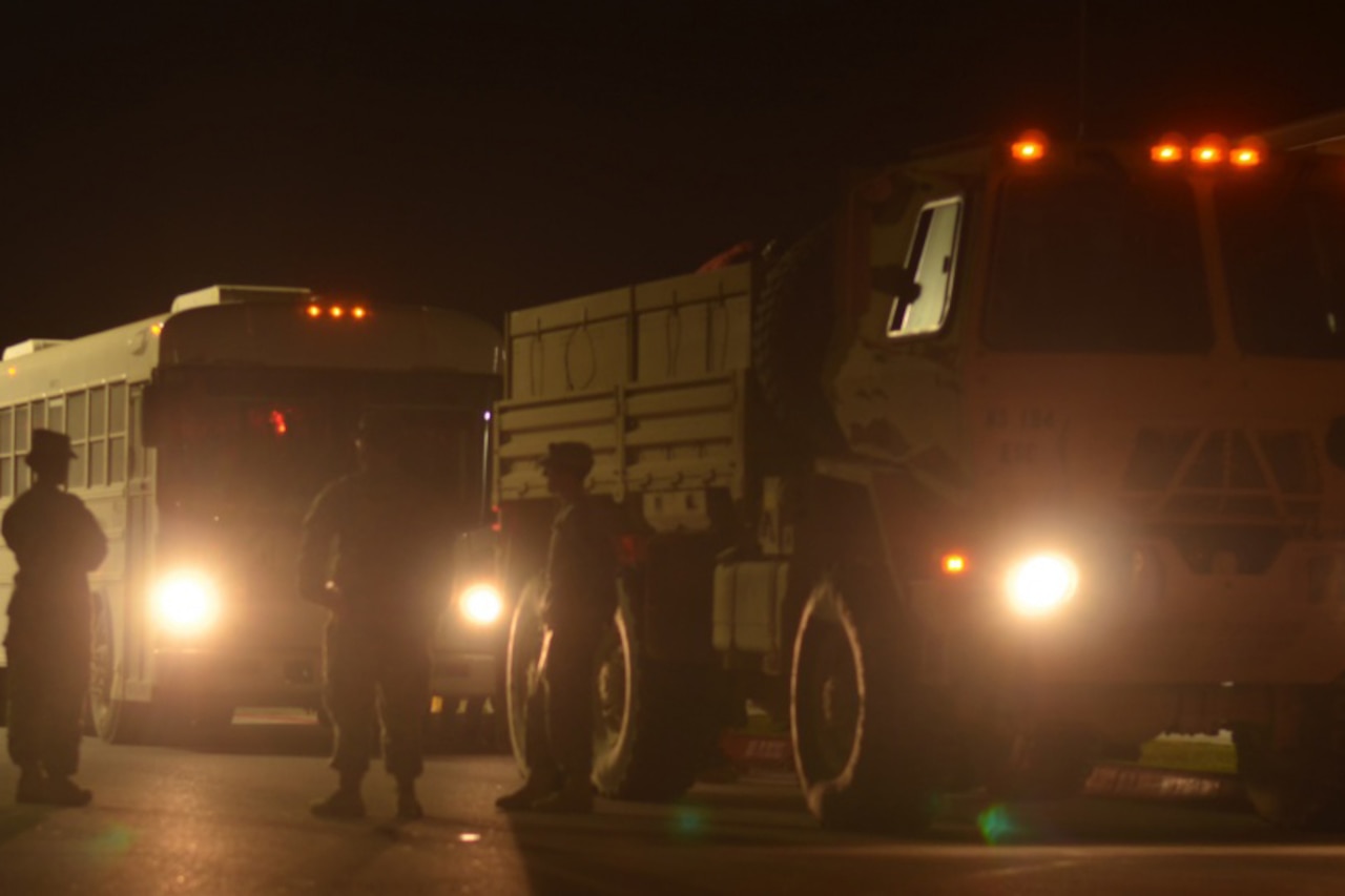 Service members stand near a bus and a military vehicle. It is dark outside.