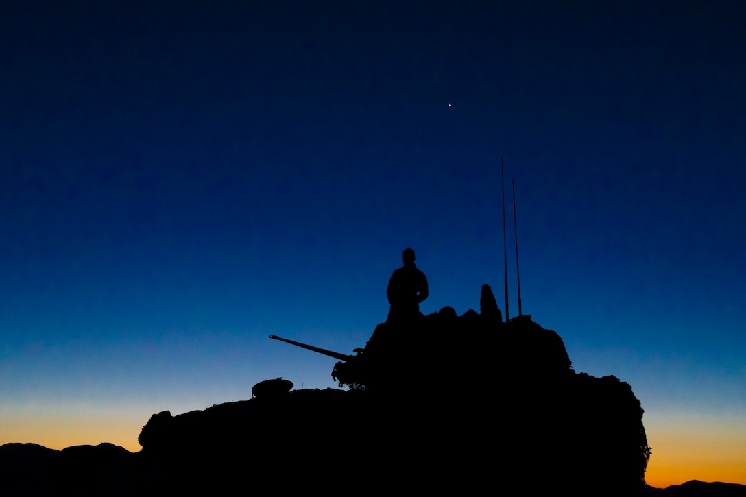 A Marine, shown in silhouette, stands atop a military vehicle with a vivid blue sky in the background