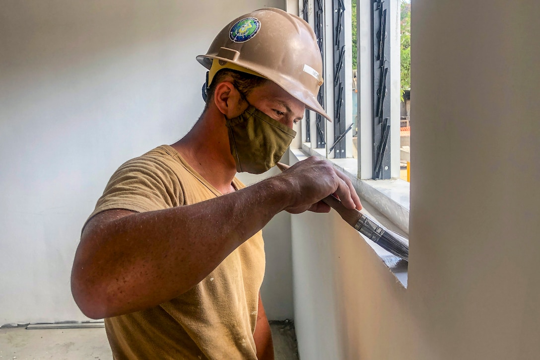 A sailor wearing a hardhat and face mask paints along a window ledge.