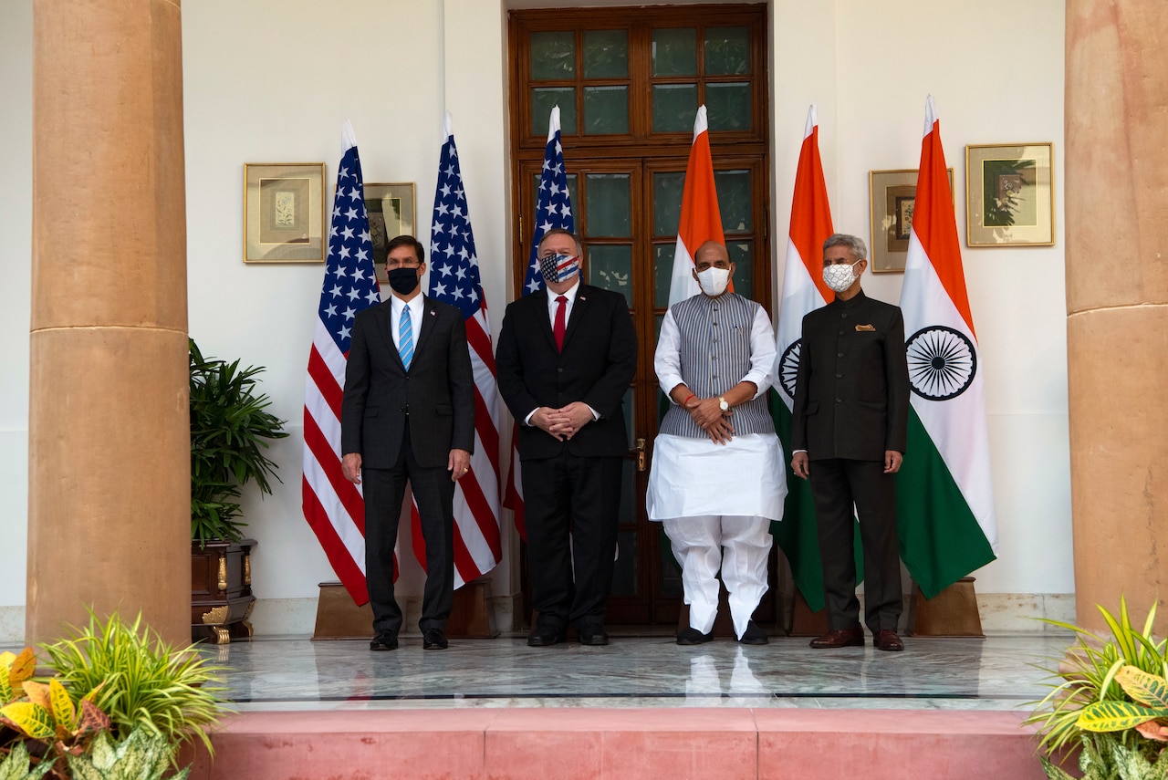 Four men wearing masks pose for a photo in front of the U.S. and Indian flags.