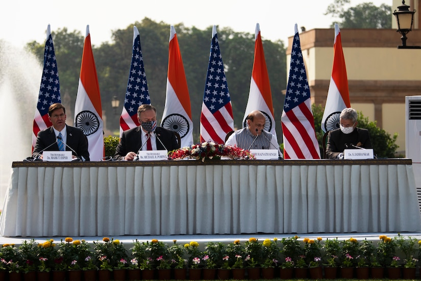 Four men sit outside at a table in front of U.S. and Indian flags.