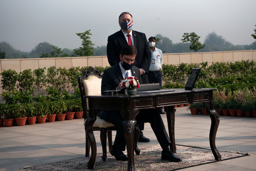 A man sits at a desk to sign a document. Another man stands behind him looking on. Another man is further in the background.