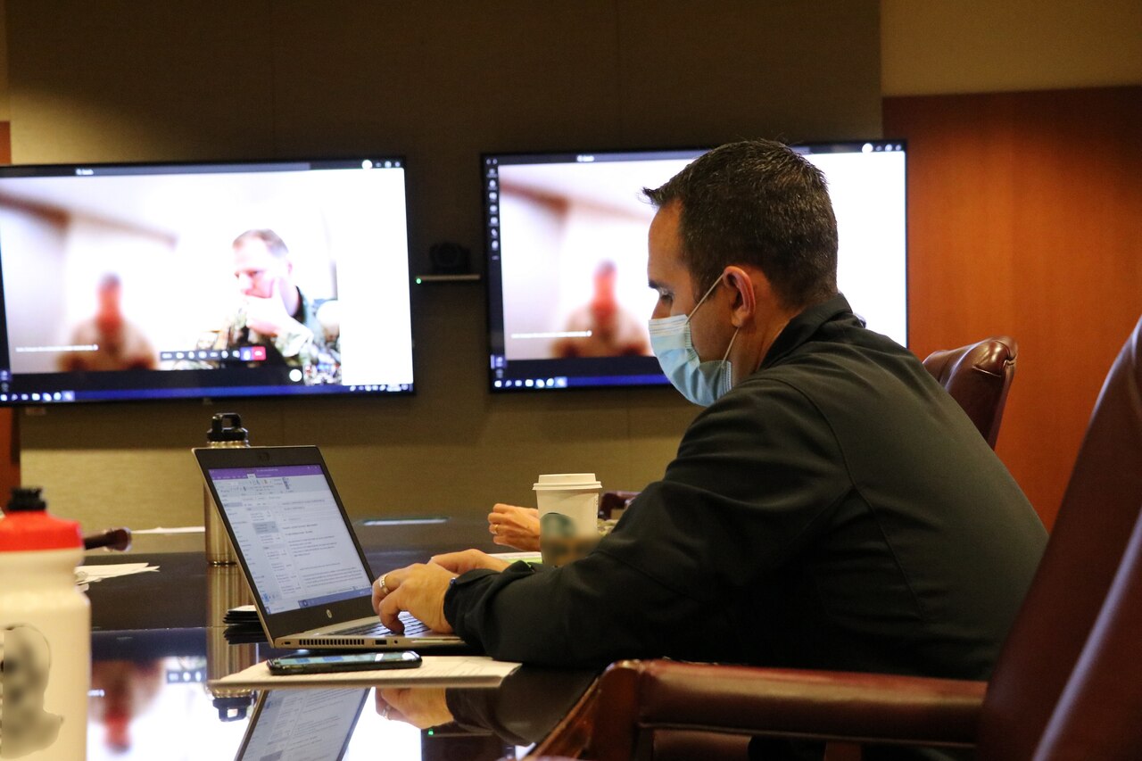 A man in a uniform is working on a laptop at a conference table. Screens in the background indicate that others are participating virtually.