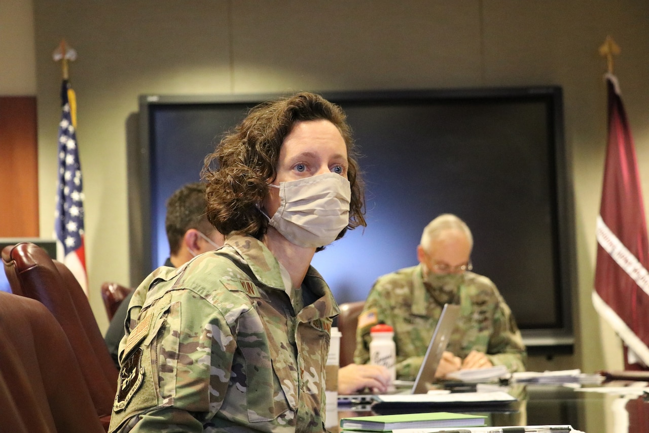 A woman in a military uniform and wearing a face mask participates in a discussion. Two other participants are in the background.
