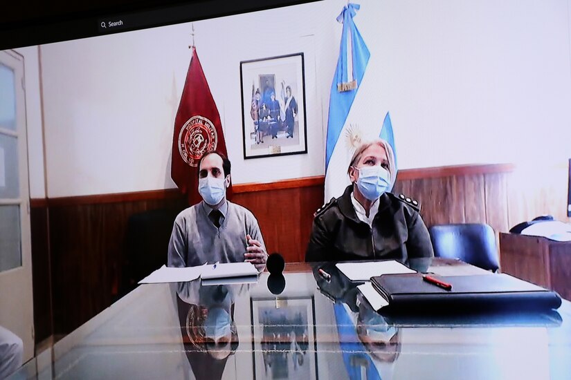 A civilian and a woman officer from a foreign army appear on a teleconference screen. Two flags from foreign entities are in the background.