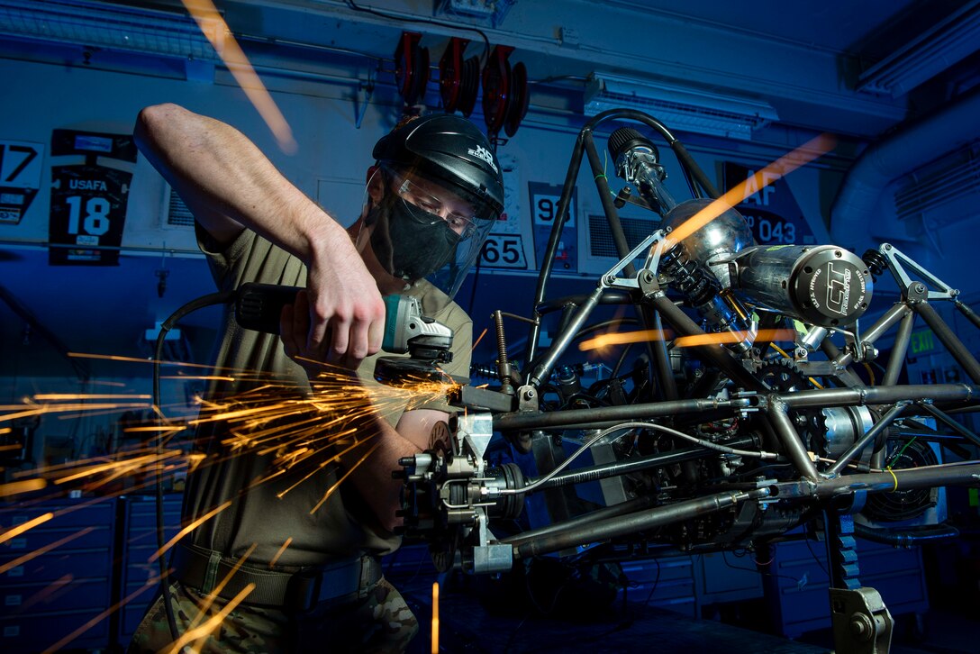 A cadet wearing protective gear uses a machine on a race car as sparks fly.