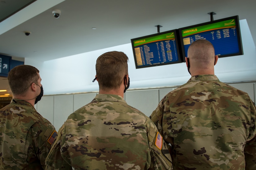 Three service members look at the electronic signs that list arriving flights at an airport.