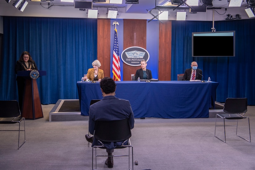 Three people sit at a long table. A woman stands at a lectern. In the foreground, a man sits in a chair, facing them.