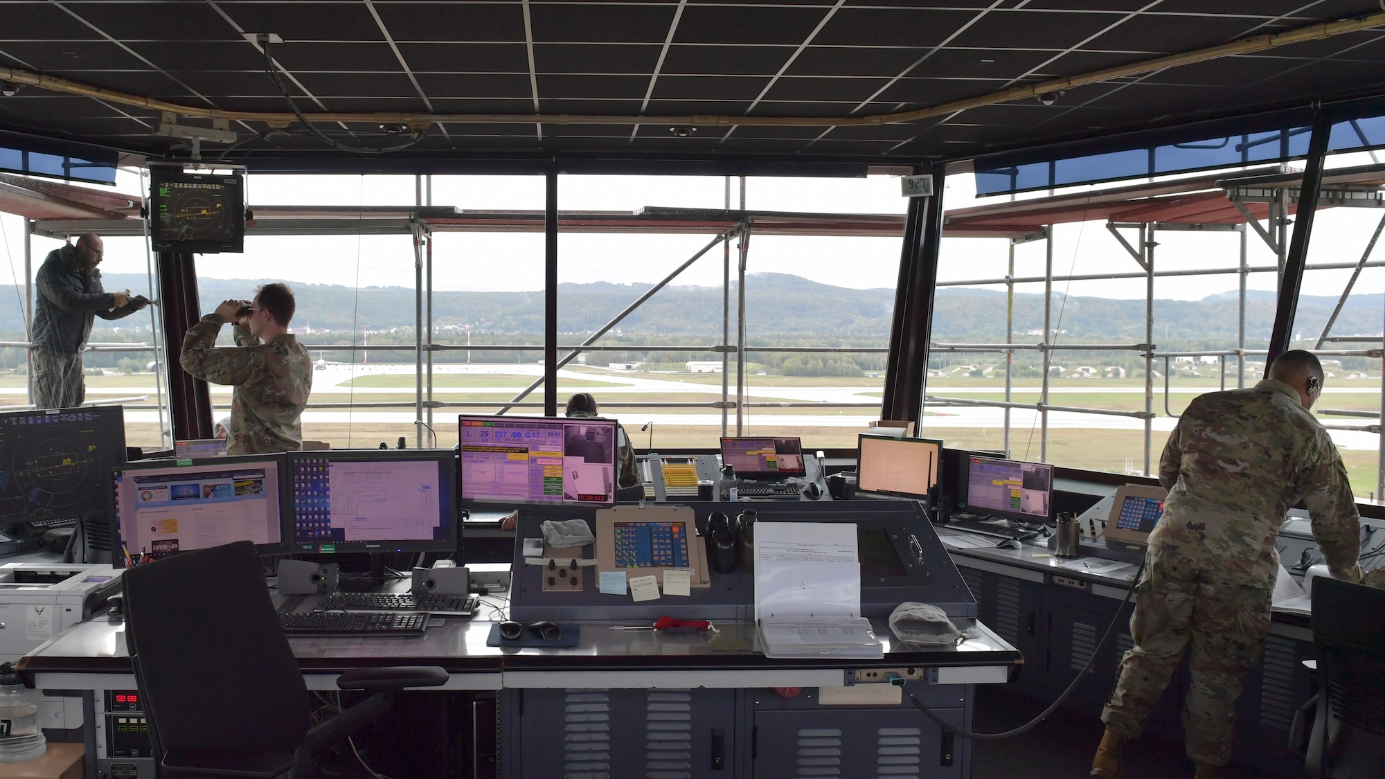 Three Airmen standing in a control tower room, and one construction worker standing outside the room.