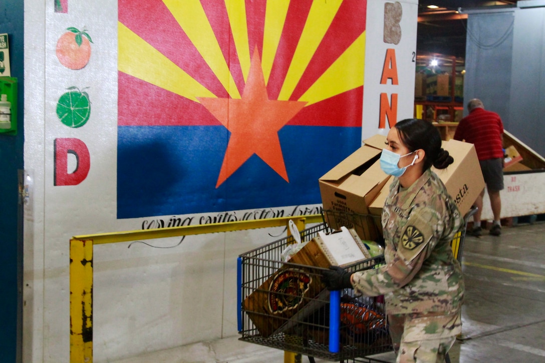 A soldier wearing a face mask and gloves pushes a cart with boxes.