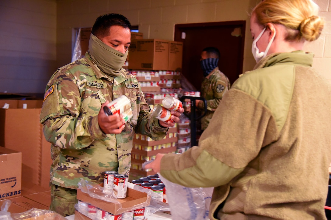 A service member prepares to put cans of food into a plastic bag held by a service member; both are wearing face masks.