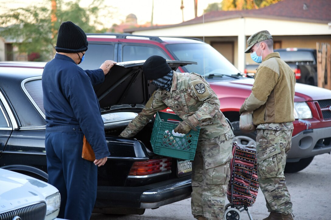 Two service members wearing face masks and gloves carry boxes of food to a vehicle; a man in civilian clothing stands next to the vehicle.