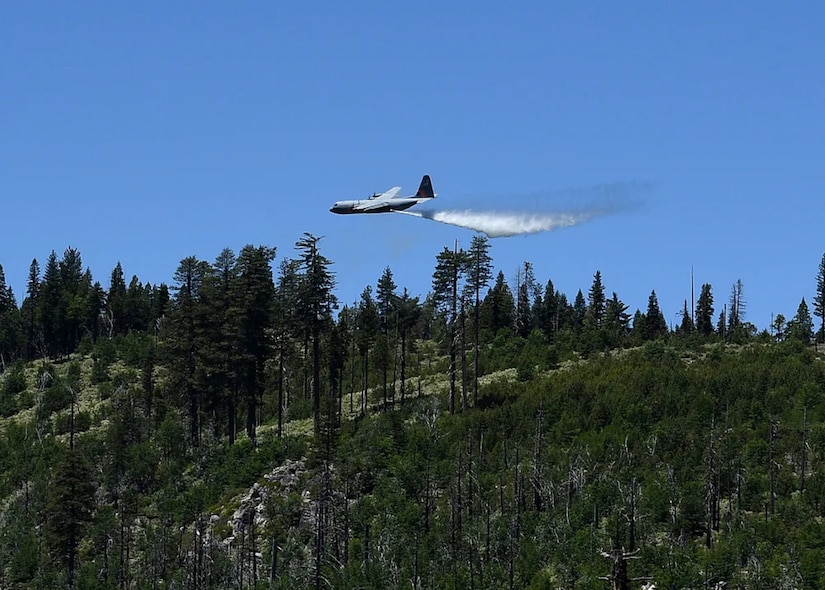 A C-130 aircraft releases water over a forest as it flies through the air.