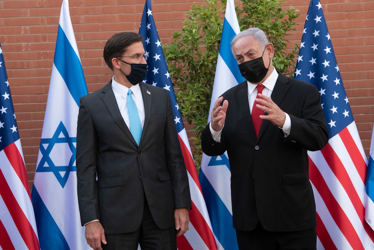 Two men stand in front of Israeli and American flags.