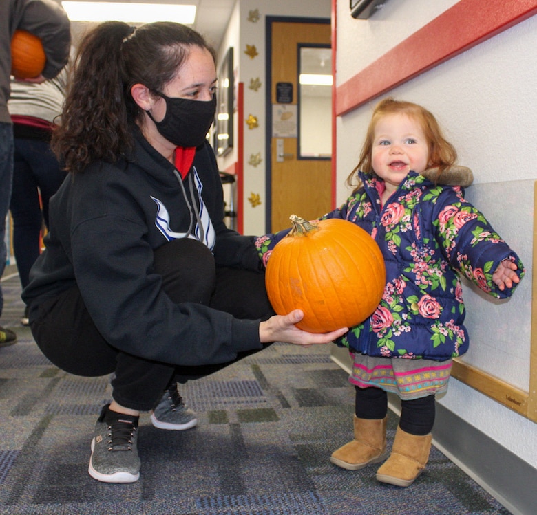 Staff Sgt. Megan Aycock, Colorado Springs Regional Command Post emergency actions controller, left, hands a pumpkin to her daughter Mabry, 1, as they prepare to leave the Child Development Center at Schriever Air Force Base Oct. 23, 2020. Parents picked up pumpkins to decorate for fun or to enter into the 50th Force Support Squadron’s pumpkin carving contest. (U.S. Space Force photo by Marcus Hill)