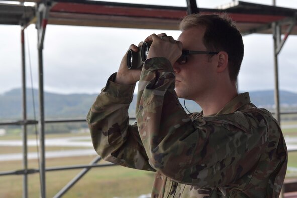 Three Airmen standing in a control tower room, and one construction worker standing outside the room.