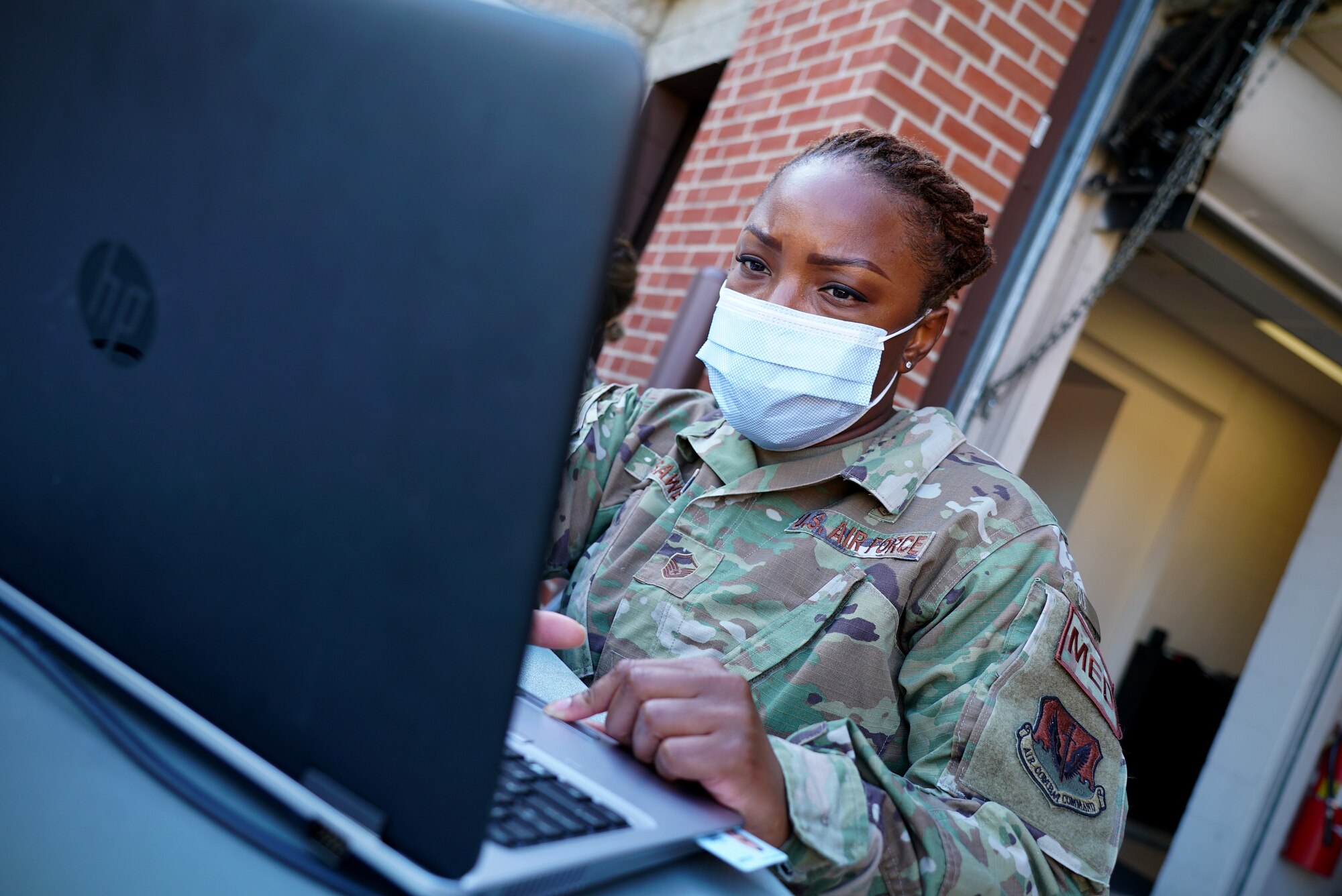A female medical technician wears a protective face mask as she sits behind a grey laptop.