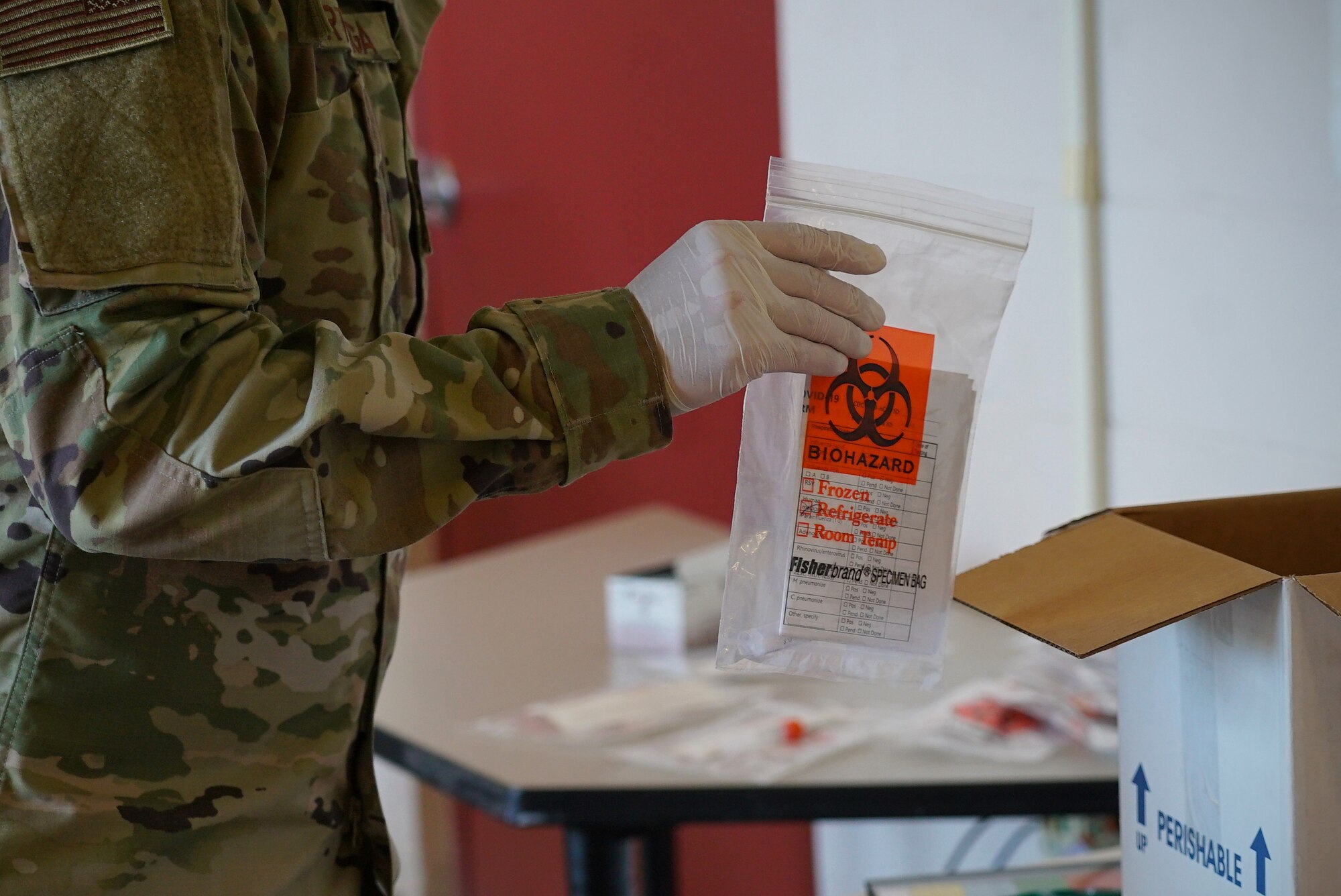 A gloved hand holds a clear and orange biohazard bag near a box to be shipped later.