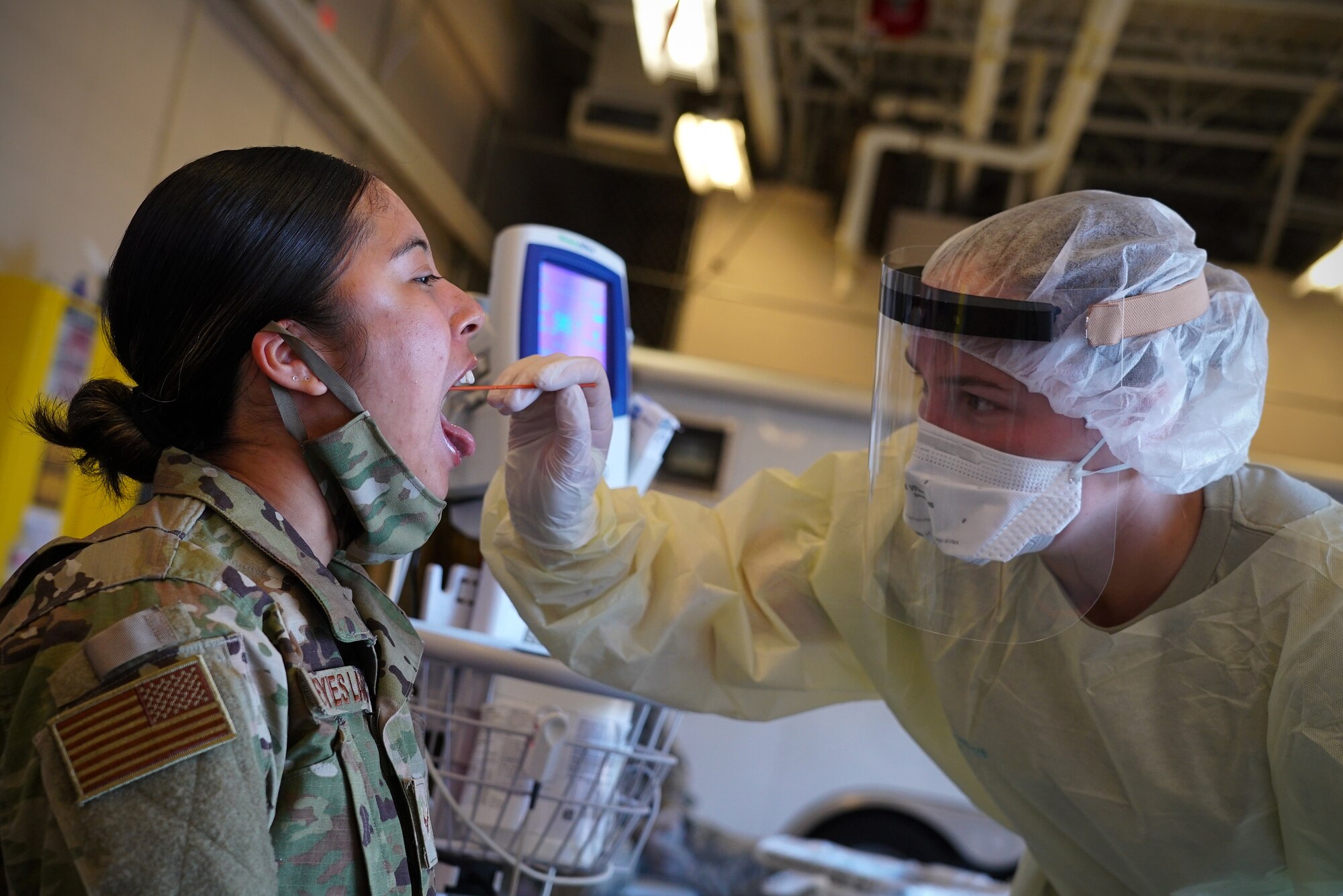 A woman in military uniform opens her mouth and sticks out her tongue as a medical technician wearing gloves, a face mask and protective gown swabs the back of her throat.