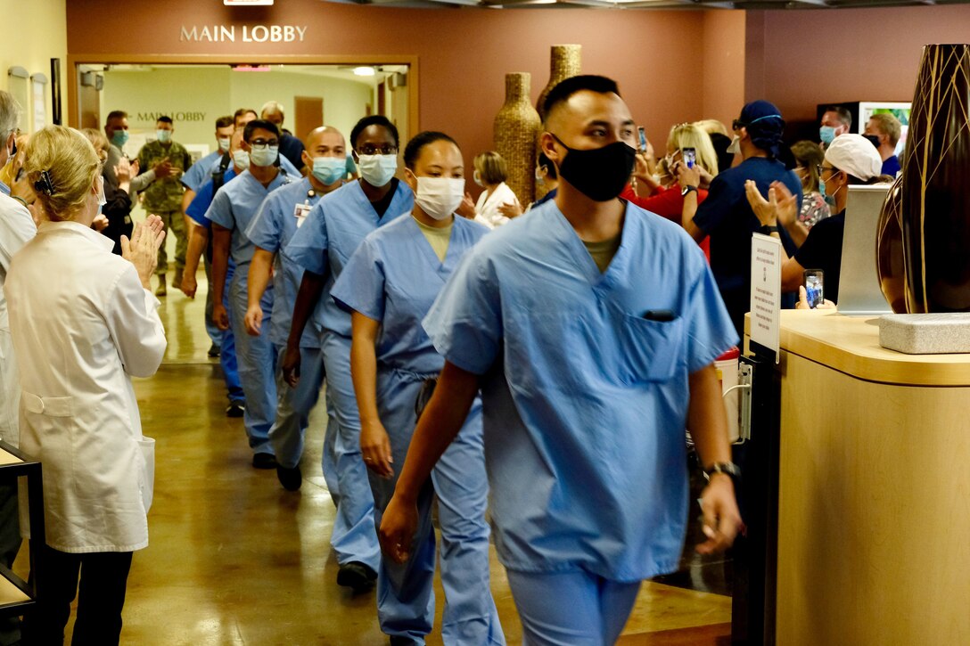 A line of airmen in medical uniforms walk down a corridor while other people clap for them.