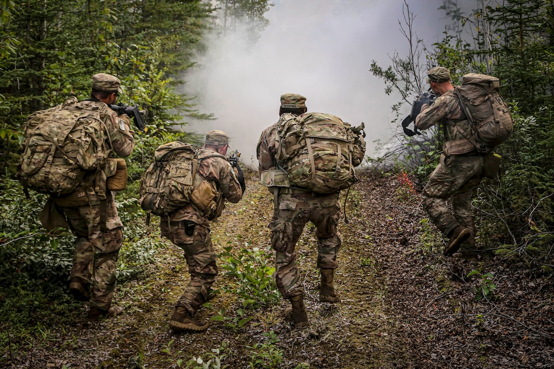 Four soldiers, shown from behind, crouch and point weapons while moving on a forest trail toward a smoke screen.