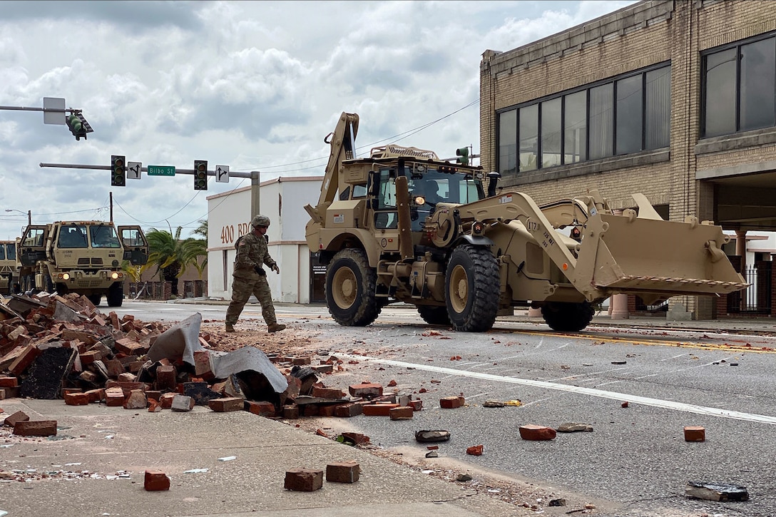 A National Guardsman walks toward a front-end loader with a stack of bricks in the foreground.