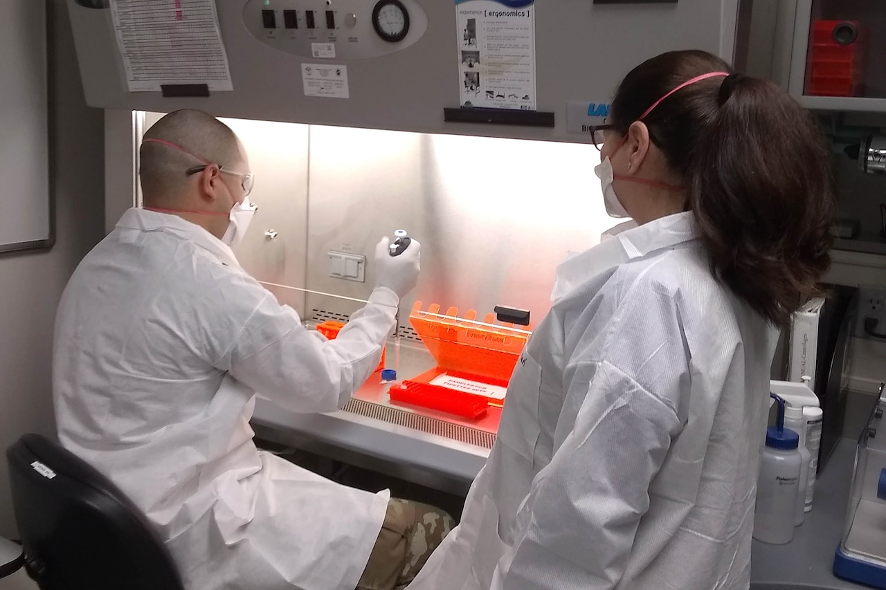 A lab technician processes a vector sample while a microbiologist observes.Both are wearing lab coats and masks.